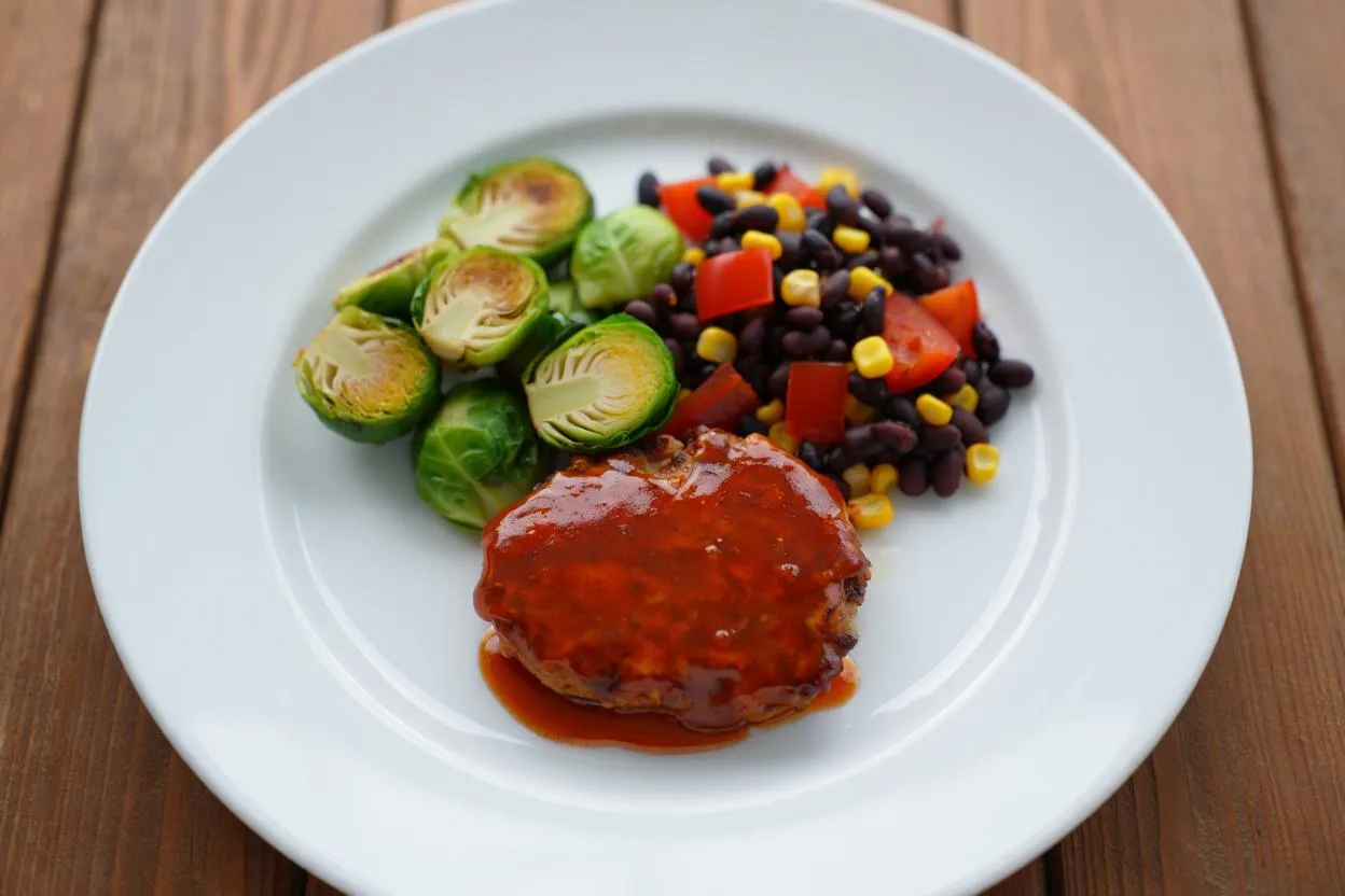 Glazed patty served with halved Brussels sprouts and a black bean, corn, and pepper salad on a white plate.
