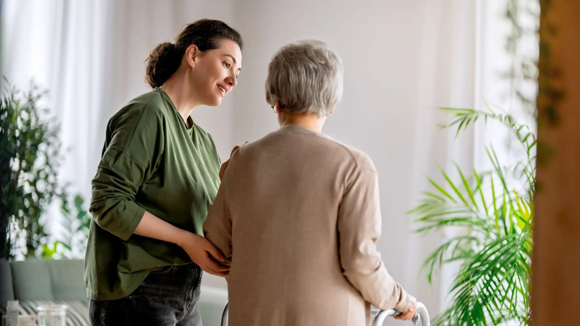 A caregiver in a green shirt assists an elderly woman using a walker.