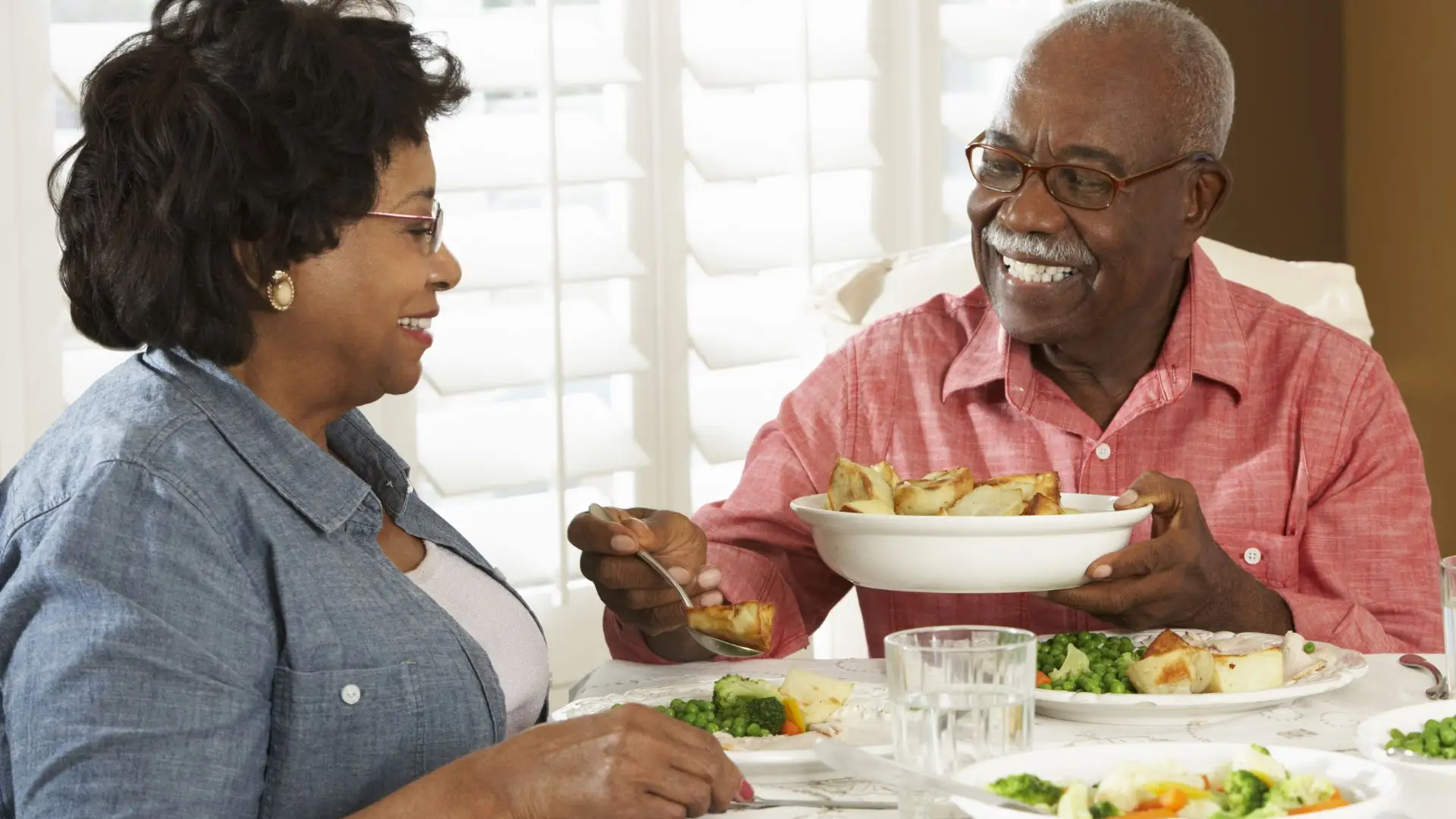 A smiling senior Black couple sitting at a dining table, sharing a meal.