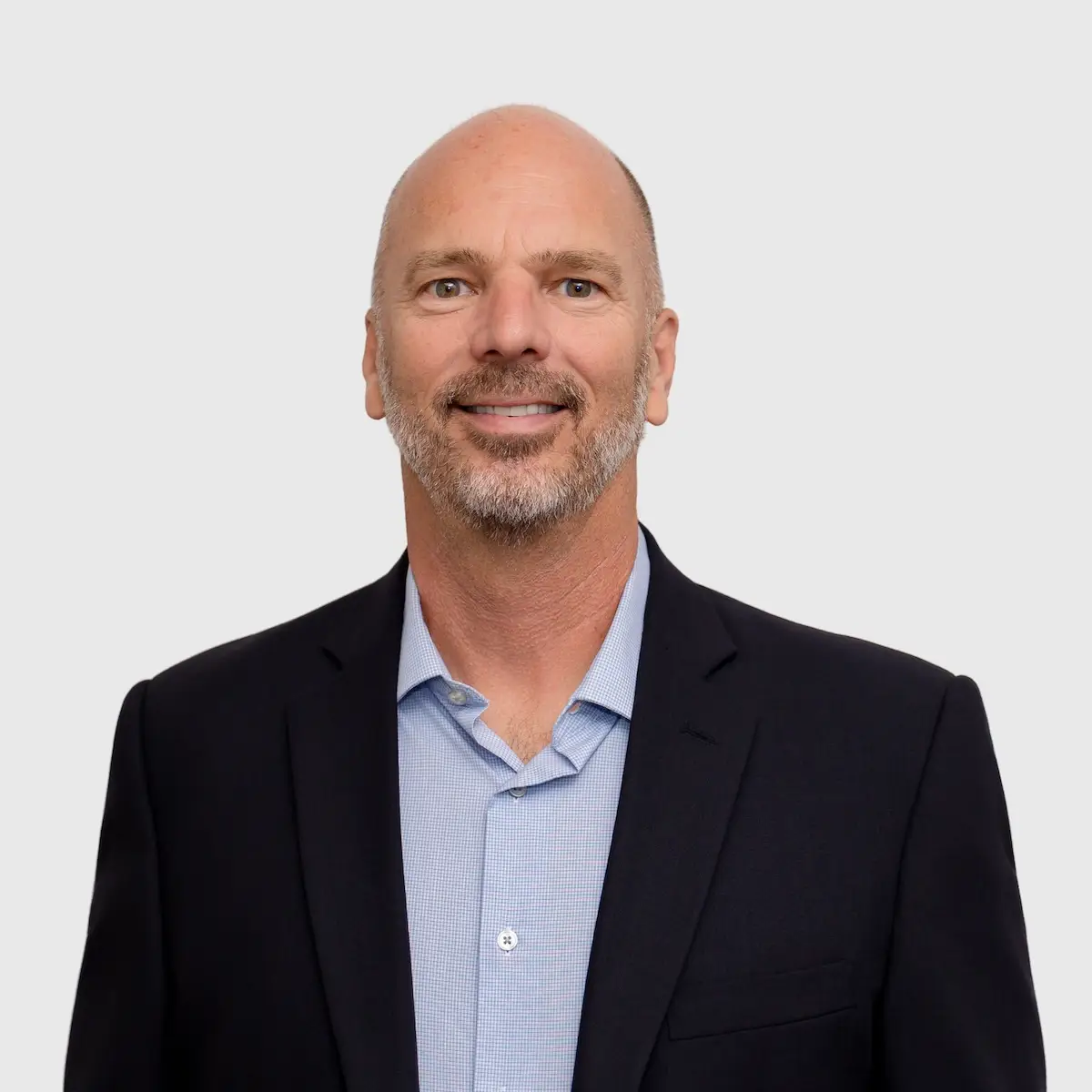 Smiling bald man with a short beard wearing a dark blazer and light blue shirt against a light gray background.