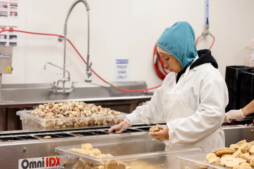 A food prep worker in a white coat and blue hood prepares meals at a commercial kitchen station.