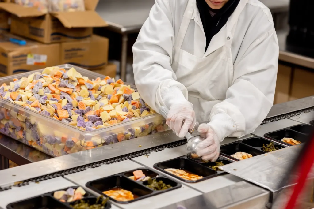 A food service worker wearing gloves packages prepared meals into black compartment trays on an assembly line.