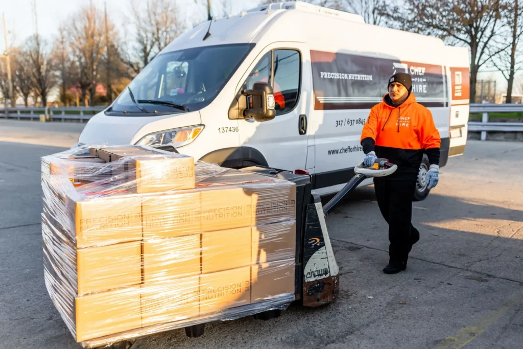 Delivery worker operating a pallet jack loaded with wrapped boxes in front of a white delivery van.