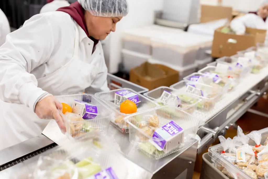 A worker in a hairnet packs plastic meal containers with fruit and milk cartons on an assembly line.